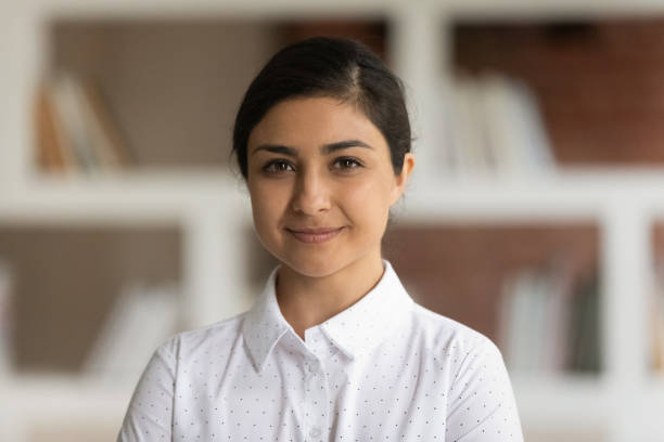 University or school teacher, business lady had shot portrait concept. Young attractive Indian ethnicity woman in white shirt pose in library room with bookshelves on background smile look at camera